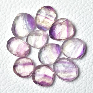 May include: A collection of ten faceted fluorite gemstones. The stones are a mix of purple, white, and clear, with a translucent appearance. The stones are oval shaped and have a smooth, polished finish.