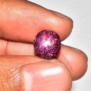 Puede incluir: Una gema redonda y pulida, de un profundo color magenta. La gema tiene un aspecto moteado con áreas más claras que reflejan la luz. La piedra se sostiene entre los dedos, mostrando su tamaño y color.