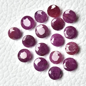 May include: A collection of 17 faceted ruby gemstones, each with a deep pink hue and a round shape. The stones are arranged in a loose cluster on a white surface.