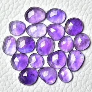 Pode incluir: Uma coleção de 16 pedras preciosas de ametista facetadas em vários tons de roxo. As pedras estão dispostas em círculo sobre um fundo branco.