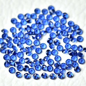 May include: A collection of small, round, faceted blue gemstones scattered on a white surface. The gemstones have a brilliant, reflective quality, with multiple facets catching the light. The overall effect is one of sparkle and depth.