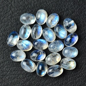 May include: A collection of oval-shaped moonstone cabochons, with a milky white color and iridescent blue flashes. The stones are arranged in a loose pile on a black surface.