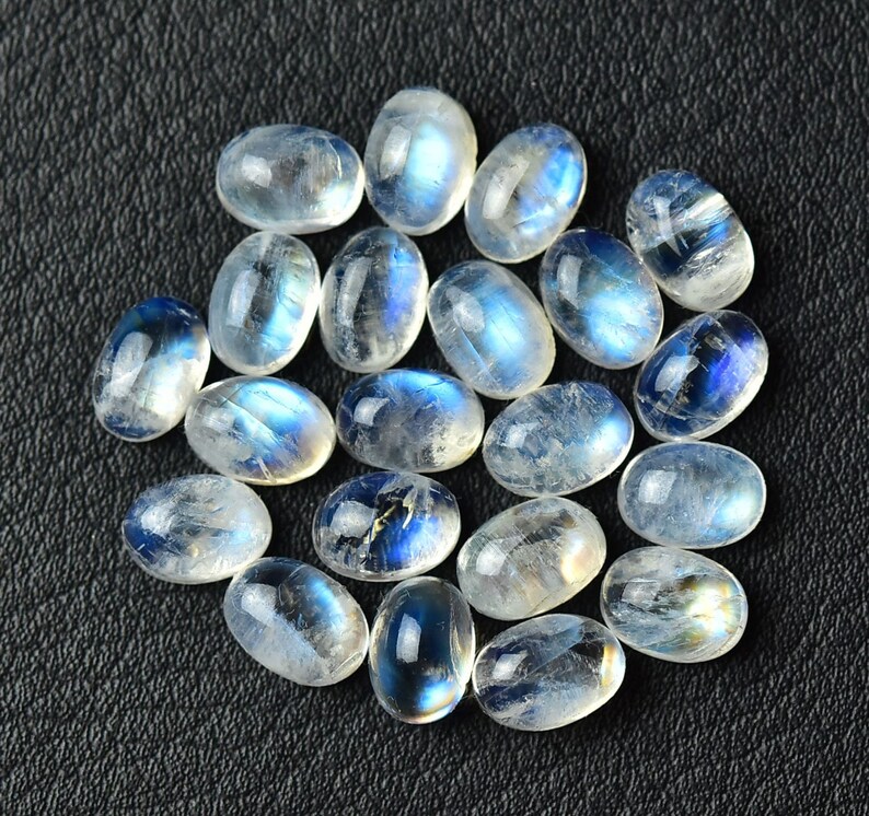 May include: A collection of oval-shaped moonstone cabochons, with a milky white color and iridescent blue flashes. The stones are arranged in a loose pile on a black surface.