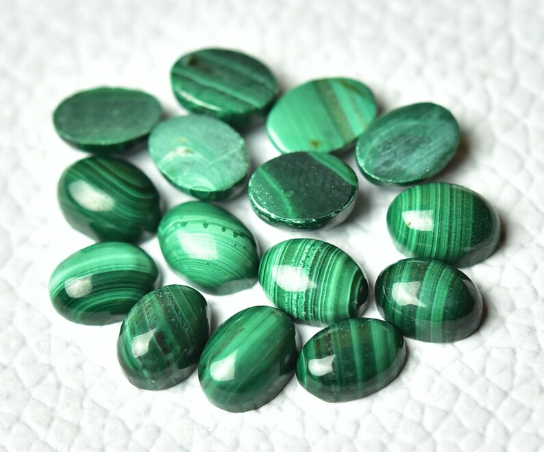 May include: A collection of 14 oval-shaped malachite cabochons in various shades of green. The stones have a smooth, polished finish and display intricate patterns of swirling green hues.