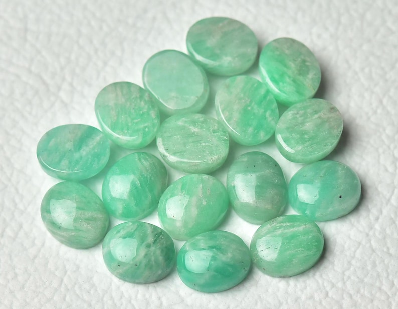 May include: A collection of 15 oval-shaped green gemstones, likely amazonite, arranged on a white surface. The stones are smooth and polished, with a variety of shades of green.