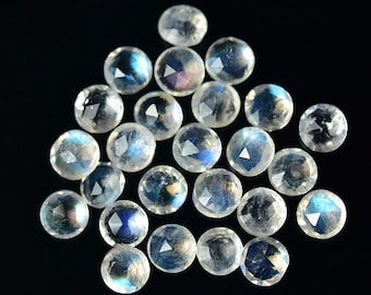 May include: A collection of round, faceted moonstone gemstones. The stones are clear with iridescent blue and white flashes, set against a black background. The gemstones are arranged in a scattered pattern.