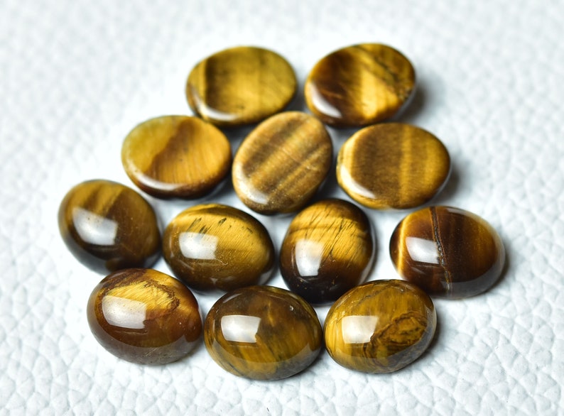 K&ouml;nnte beinhalten: Eine Sammlung von 12 ovalen Tigeraugen-Edelsteinen. Die Steine sind eine Mischung aus goldbraunen und braunen Farben mit einem chatoyanten Effekt.