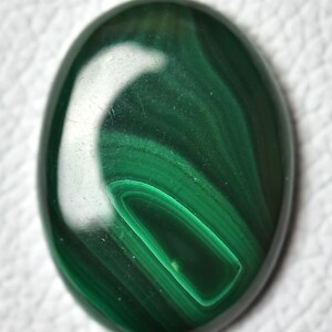 May include: A smooth, oval-shaped malachite cabochon with a deep green color and swirling patterns. The stone has a polished finish and is set against a white background.
