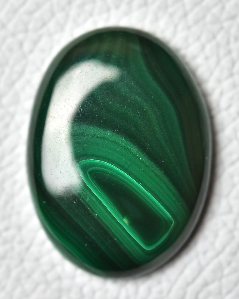 May include: A smooth, oval-shaped malachite cabochon with a deep green color and swirling patterns. The stone has a polished finish and is set against a white background.