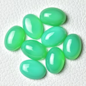 Könnte beinhalten: Acht ovale, polierte Edelsteine in verschiedenen Grüntönen liegen auf einer weißen, strukturierten Oberfläche. Die glatten, glänzenden Steine reflektieren das Licht und zeigen ihre lebendige Farbe.