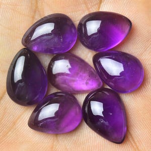Pode incluir: Sete pedras preciosas de ametista polidas em forma de gota. As pedras variam em tons de roxo, do violeta escuro ao lavanda mais claro. As gemas são lisas e refletem a luz, sugerindo um alto polimento. A disposição está em uma superfície clara.