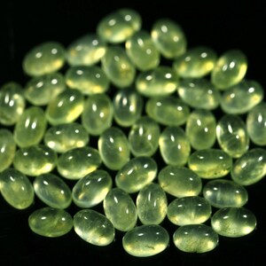 May include: A collection of smooth, oval-shaped green gemstones, likely peridot, arranged on a black background.