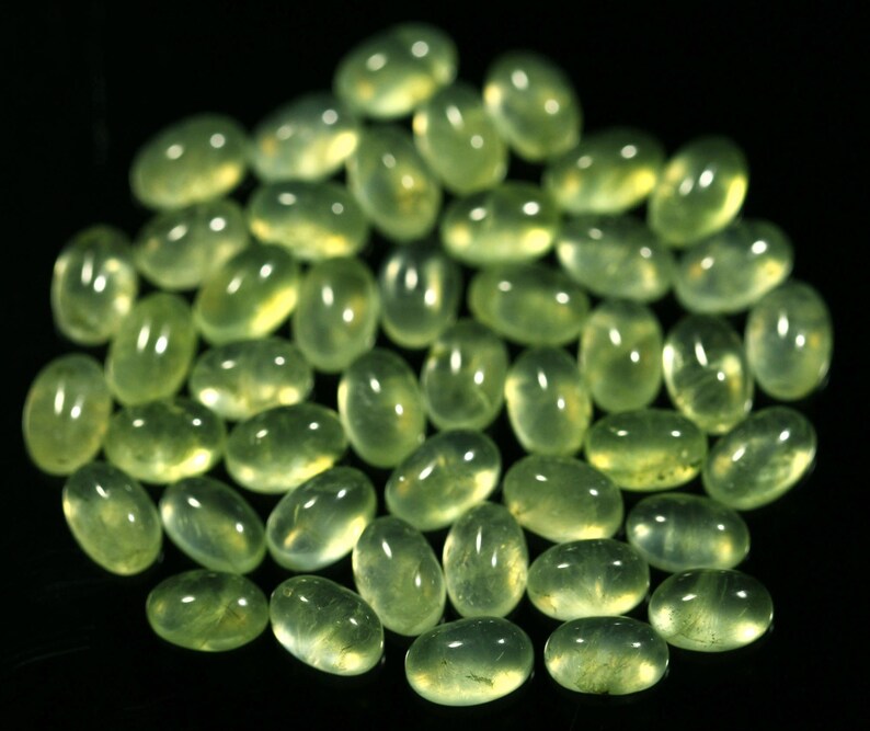 May include: A collection of smooth, oval-shaped green gemstones, likely peridot, arranged on a black background.