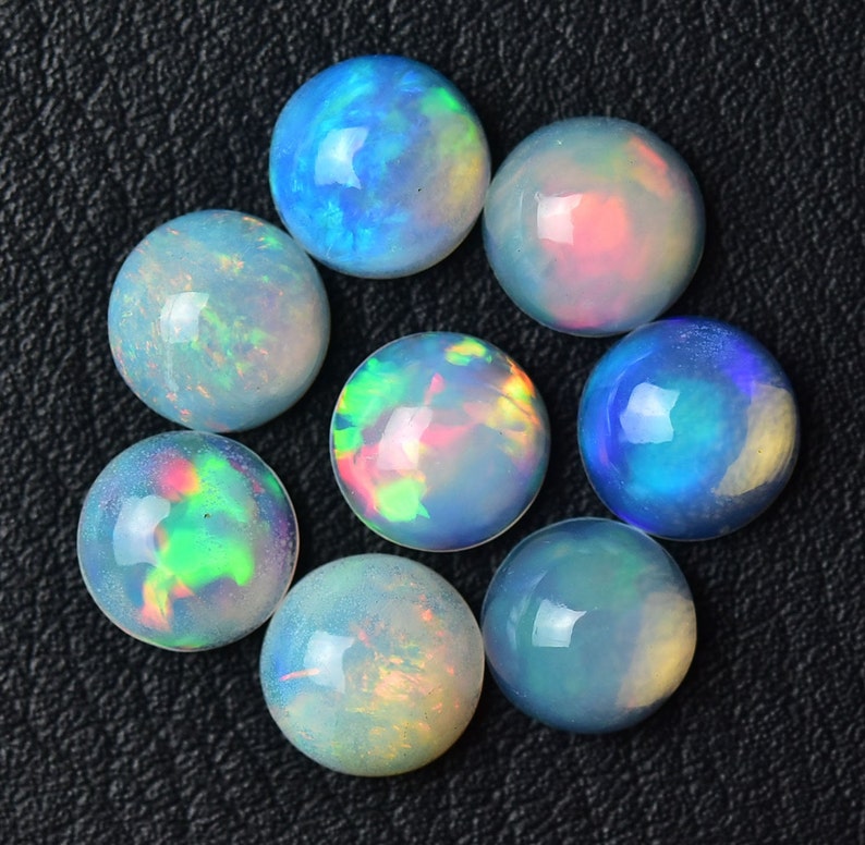 Pode incluir: Sete caboch&otilde;es de opala redondos em v&aacute;rios tons de azul, verde e rosa. As opalas est&atilde;o dispostas em um c&iacute;rculo sobre um fundo preto.