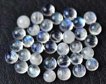 May include: A collection of round, white moonstone cabochons with iridescent blue and green flashes. The stones are smooth and polished, and they are arranged in a loose pile.