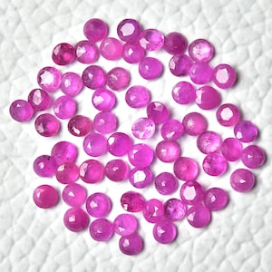 May include: A collection of pink ruby gemstones, faceted and polished, with a round shape. The gemstones are scattered on a white surface.