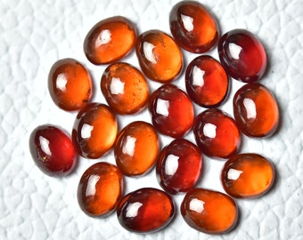 May include: A collection of 18 oval-shaped, polished, red-brown gemstones. The stones are arranged in a circular pattern on a white surface.