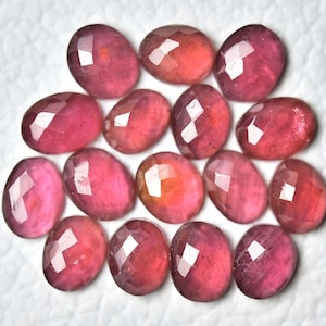Könnte beinhalten: Eine Sammlung ovalförmiger, facettierter Edelsteine in Rosa- und Rottönen. Die Edelsteine sind auf einer weißen Oberfläche angeordnet, wodurch ihre polierten Oberflächen und ihre innere Klarheit hervorgehoben werden. Die Farbtiefe der Edelsteine variiert.