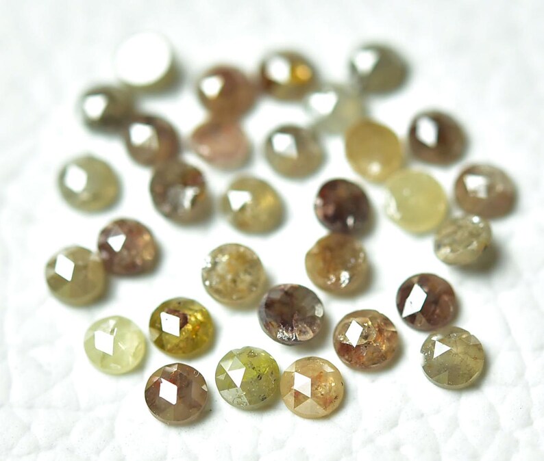 Op de afbeelding: Een verzameling ronde, gefacetteerde, bruine, gele en groene ruwe diamanten. De diamanten zijn verspreid over een wit oppervlak.