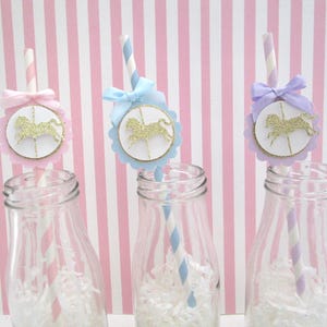 May include: Three glass bottles with white paper shreds inside. Each bottle has a striped paper straw with a round paper topper featuring a gold glitter carousel horse design. The toppers are decorated with a pink, blue, and purple ribbon.
