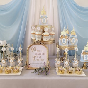 May include: A fairytale-themed dessert table with a light blue and gold color scheme. The table features cupcakes, gift boxes, and crown-shaped treats. A sign reads "Once upon a time". Decorative flowers and a light blue backdrop complete the scene.