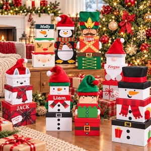 May include: A collection of stacked decorative Christmas gift boxes featuring various character designs including a penguin, nutcracker, snowman, elf, and Santa Claus. Each box set is topped with a plush hat and personalised with names.