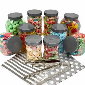 May include: Nine glass jars filled with colourful sweets, including green, red, blue, yellow, and orange sweets. The jars have grey lids and are arranged on a white and grey striped surface with two silver tongs.
