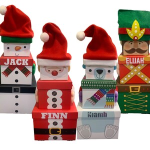May include: Four stacked Christmas gift boxes with different character themes. The boxes are decorated with red, green, white, and brown colours. The top boxes feature a snowman, Santa Claus, a polar bear, and a gingerbread man. The boxes are labelled "Jack", "Finn", "Niamh", and "Elijah".