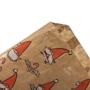 K&ouml;nnte beinhalten: Eine braune Papiert&uuml;te mit einem wiederkehrenden Muster aus Weihnachtsmannfiguren mit roten M&uuml;tzen, Zuckerstangen und dem Text "NAUGHTY OR NICE", "JOY TO ALL" sowie "HO! HO! HO!". Das Papier hat eine texturierte, nat&uuml;rlich braune Oberfl&auml;che.