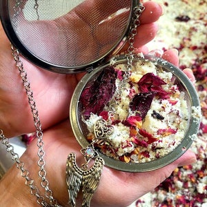 May include: A silver tea infuser with a chain and a decorative angel wing charm. The infuser is filled with dried rose petals and white powder.