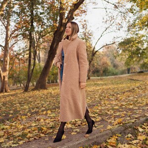 May include: A woman wearing a long, light brown fuzzy coat walks through a park covered in fallen autumn leaves.