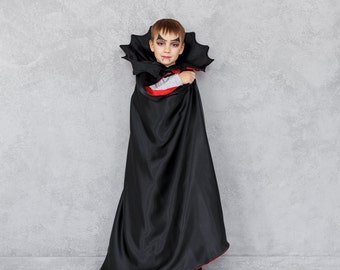 May include: A young child dressed as a vampire for Halloween. They are wearing a black cape with a red lining and have vampire makeup on their face.