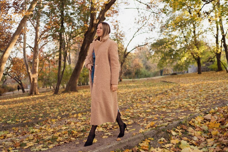May include: A woman wearing a long, light brown fuzzy coat walks through a park covered in fallen autumn leaves.
