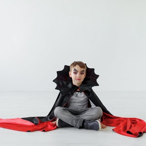 May include: A young child dressed as a vampire for Halloween. They are wearing a black cape with a red lining and have vampire makeup on their face. They are sitting on a white floor.