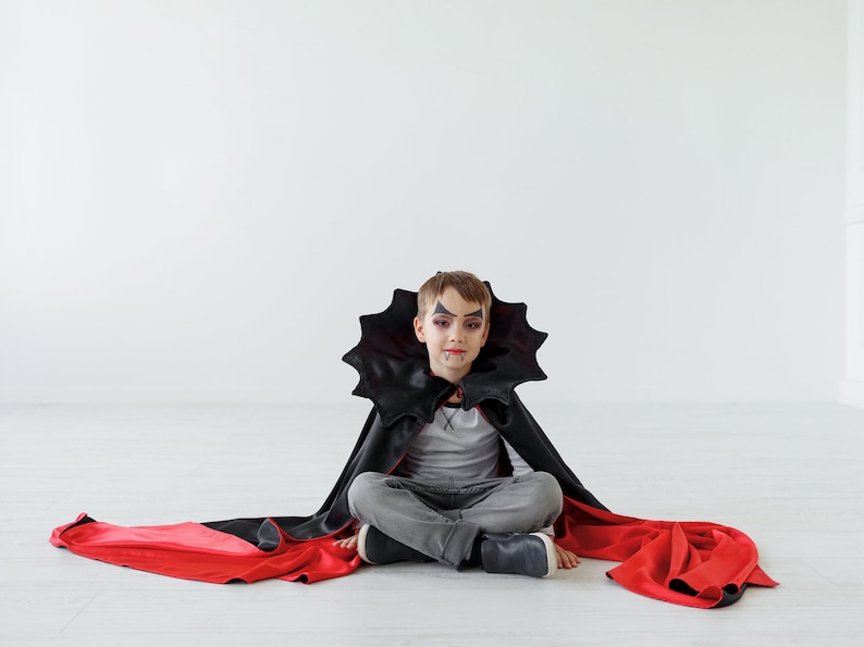 May include: A young child dressed as a vampire for Halloween. They are wearing a black cape with a red lining and have vampire makeup on their face. They are sitting on a white floor.