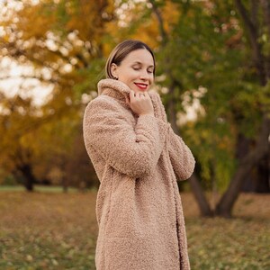 May include: A woman wearing a light brown fuzzy coat with her eyes closed. She is standing in a park with fall foliage in the background.