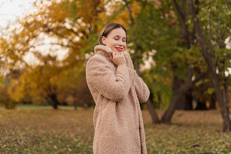 May include: A woman wearing a light brown fuzzy coat with her eyes closed. She is standing in a park with fall foliage in the background.