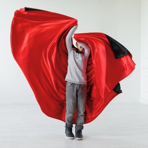 May include: A young person wearing a grey shirt and jeans is holding a red and black cape and smiling.