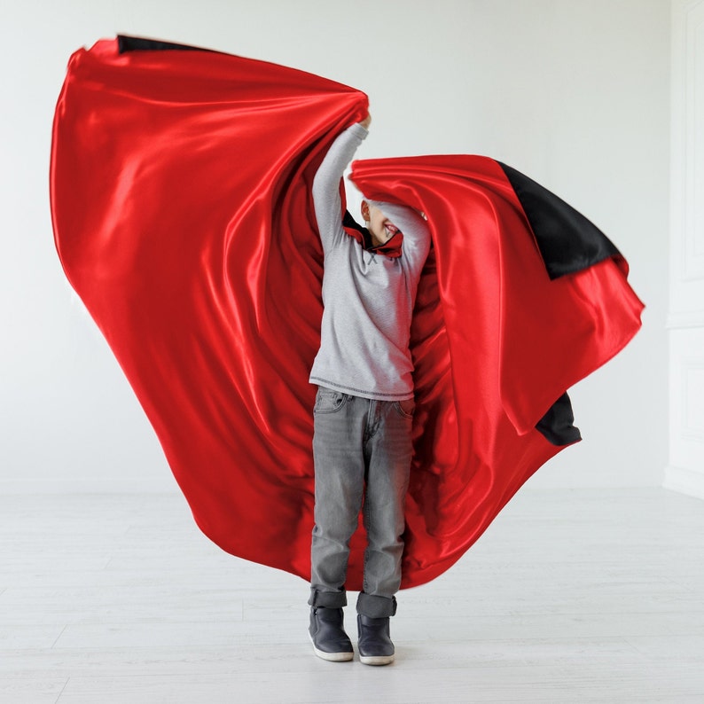 May include: A young person wearing a grey shirt and jeans is holding a red and black cape and smiling.