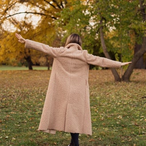 May include: A person wearing a long, light brown coat with their arms outstretched, standing in a park with fallen leaves on the ground.