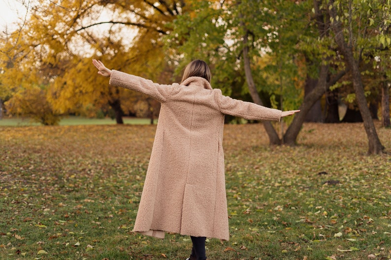 May include: A person wearing a long, light brown coat with their arms outstretched, standing in a park with fallen leaves on the ground.