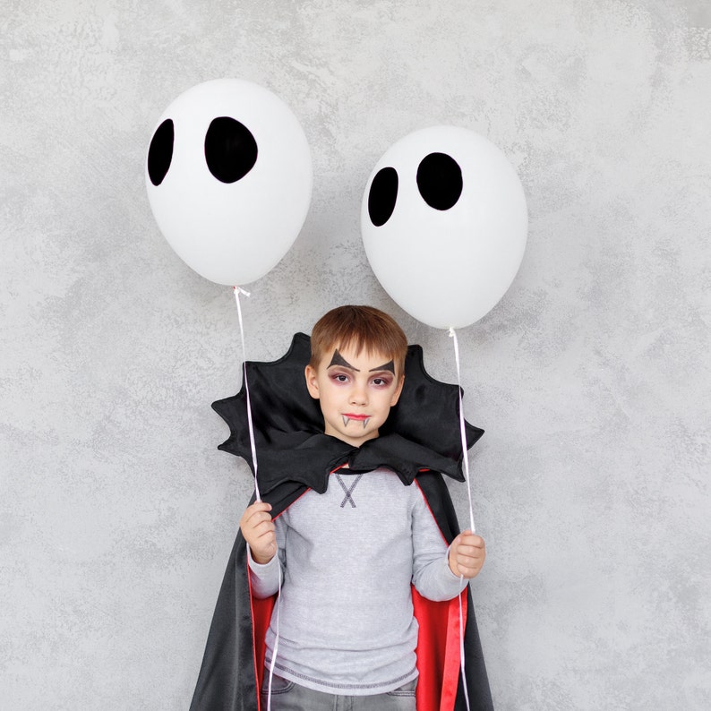 May include: A child dressed as a vampire for Halloween holds two white balloons with black eyes. The child is wearing a black cape with red lining and has vampire makeup on.