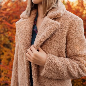 May include: A close-up of a woman wearing a light brown faux fur coat. The coat has a soft, fluffy texture and a classic collar.