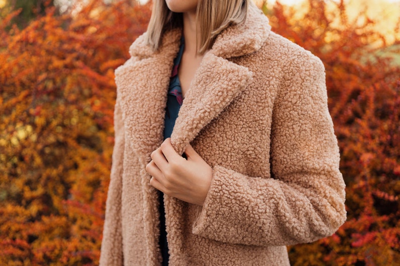 May include: A close-up of a woman wearing a light brown faux fur coat. The coat has a soft, fluffy texture and a classic collar.