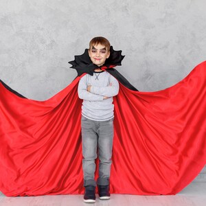 May include: A young boy dressed as a vampire for Halloween. He is wearing a red cape with a black collar and a grey shirt. He has fake vampire teeth and makeup on his face. He is standing in front of a grey wall with his arms crossed.