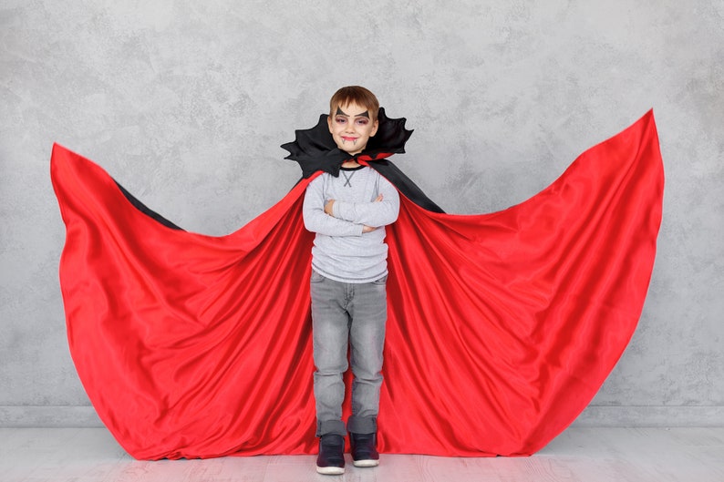 May include: A young boy dressed as a vampire for Halloween. He is wearing a red cape with a black collar and a grey shirt. He has fake vampire teeth and makeup on his face. He is standing in front of a grey wall with his arms crossed.