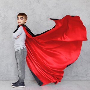 May include: A young person dressed as a vampire with a red cape. The cape is flowing in the air. The person is wearing a grey shirt, blue jeans, and black shoes. They have black makeup around their eyes and a fake fang.