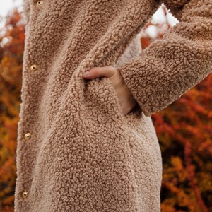 May include: A close-up of a brown faux fur coat with gold buttons. The coat is worn by a person with their hand in a pocket.