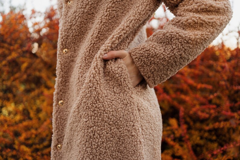 May include: A close-up of a brown faux fur coat with gold buttons. The coat is worn by a person with their hand in a pocket.