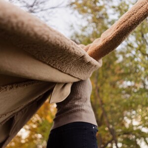May include: A person wearing a brown fuzzy coat with the sleeves spread out, standing in a forest with fall foliage.